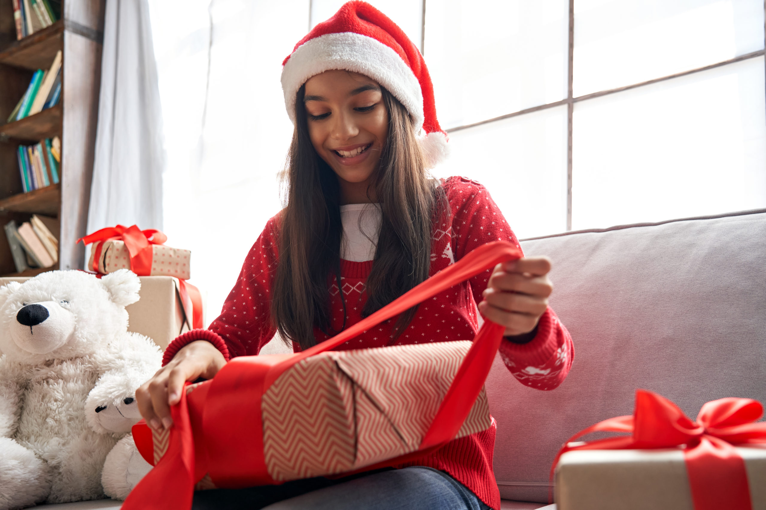Happy indian kid girl unties red ribbon opens Christmas gift box at home. Smiling cute latin child wearing santa hat receiving xmas New Year present sitting on couch celebrating winter holidays.