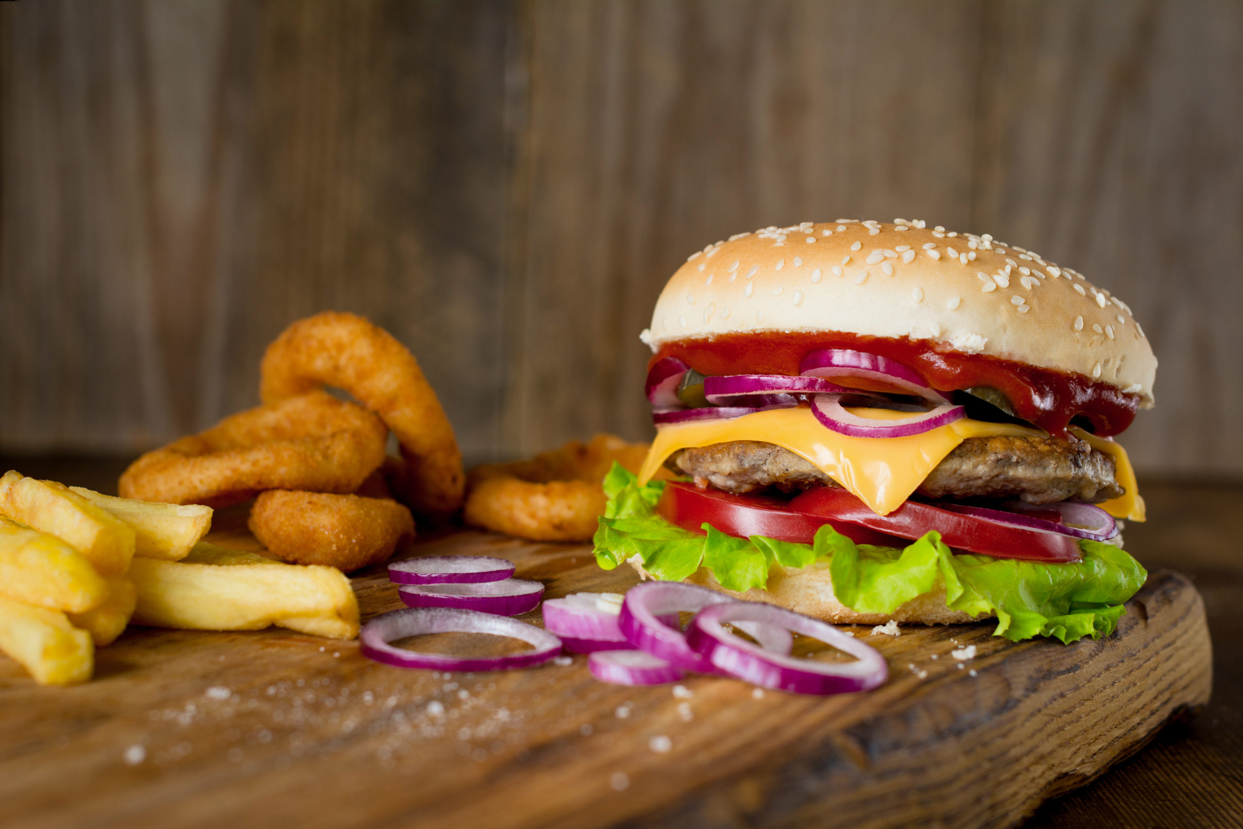 Cheeseburger, french fries and onion rings on wooden chopping board over wooden backdrop