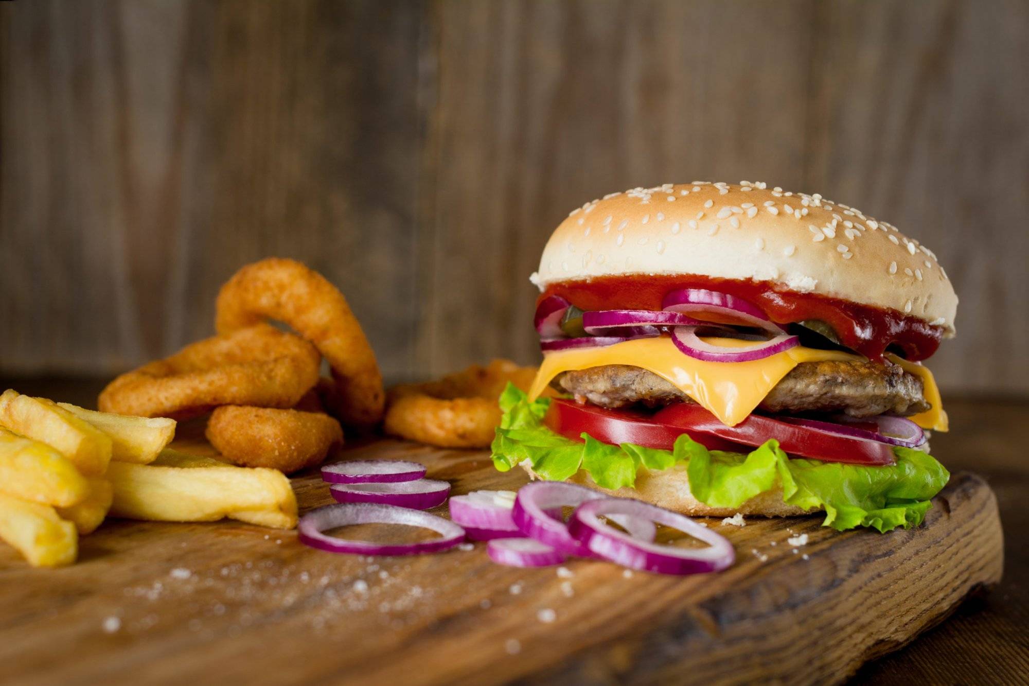 Cheeseburger, french fries and onion rings on wooden chopping board over wooden backdrop Cheeseburger, french fries and onion rings on wooden chopping board over wooden backdrop