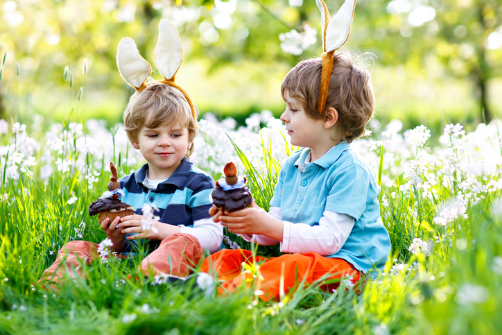 Two little boy friends in Easter bunny ears eating chocolate cakes and muffins